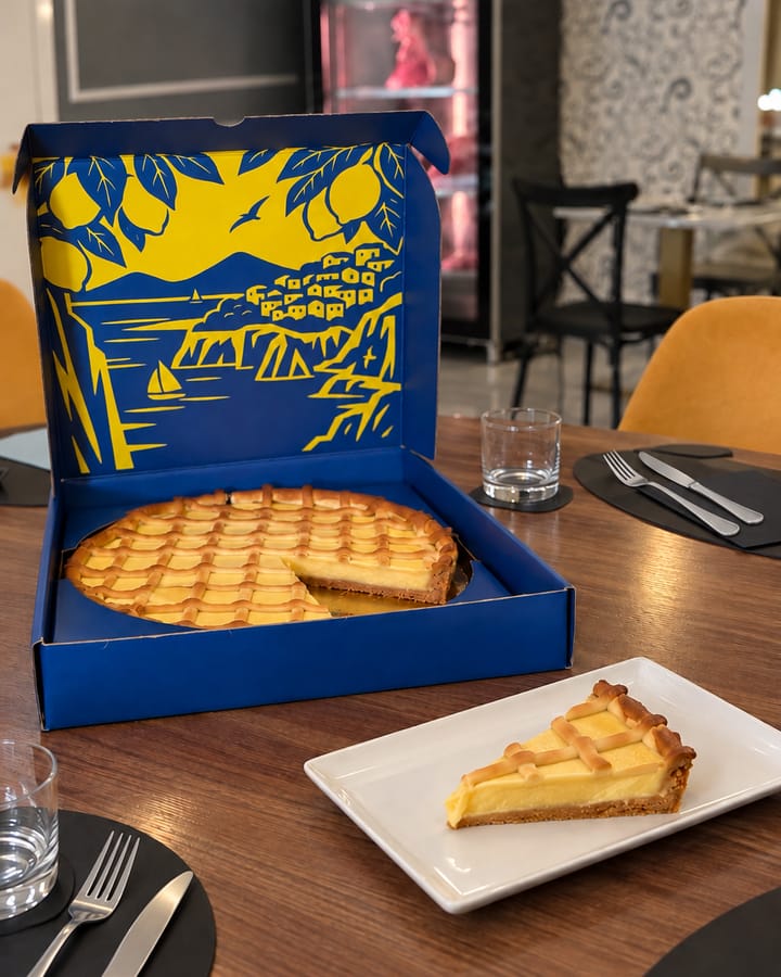 La crostata nella sua scatola blu e gialla, con una fetta servita nel piatto su un tavolo di Rovente